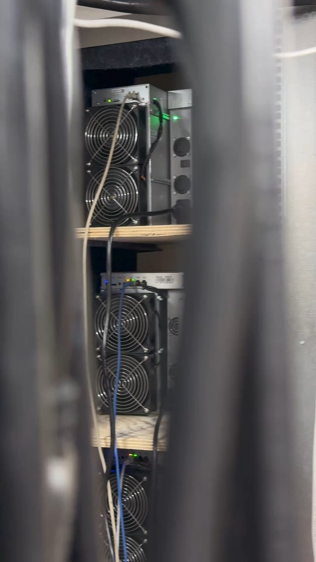 A stack of Bitcoin mining rigs with cooling fans and indicator lights sits on wooden shelves, with various cables connecting the units. The image illustrates the post's point that residential electricity rates can make home mining operations costly and inefficient.