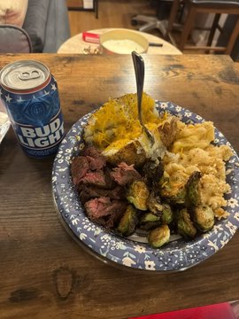 A meal featuring steak, mac and cheese, Brussels sprouts, and mashed potatoes on a floral-patterned plate sits next to a can of Bud Light on a wooden table, visually tying into the "DOGUST + BEER = BUDDOG" caption by pairing hearty comfort food with a classic beer.