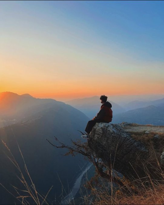A person sits on the edge of a rocky cliff at sunset, gazing out over a vast mountain valley bathed in warm orange light.
