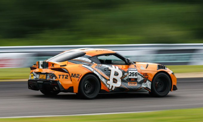 An orange sports car with a Bitcoin logo and race number 256 speeds along a racetrack with motion-blurred greenery in the background.