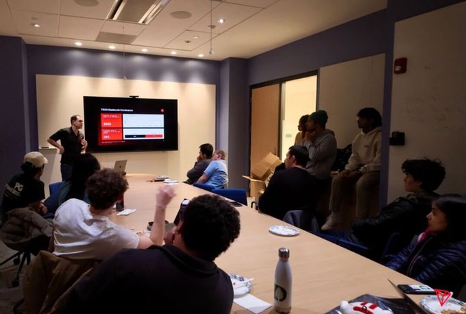 A group of people sitting around a conference table in a meeting room, with a presentation displayed on a screen at the front.