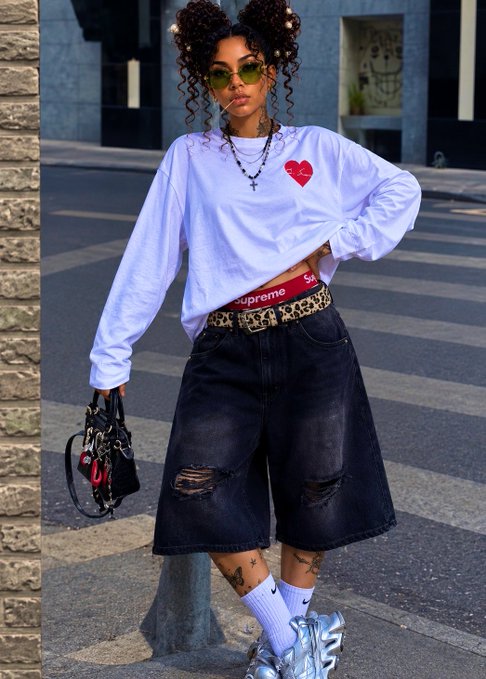 A woman poses in streetwear fashion wearing a white cropped long-sleeve shirt with a red heart, dark denim jorts with a leopard print belt, white Nike socks, and chunky silver sneakers, holding a small black bag.