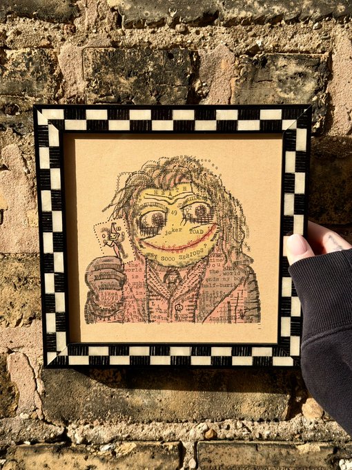 A hand holds a small black-and-white checkered picture frame against a brick wall, displaying a portrait drawing of a person made from dictionary text and coffee stains.