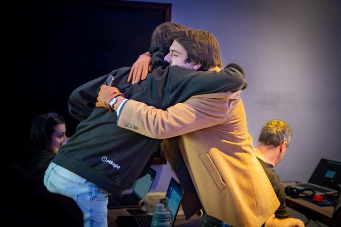 Two men share an emotional hug, one of whom is wearing a dark sweatshirt with the "midnight" logo visible on the back. The image captures a moment of celebration, likely reflecting the pride and success mentioned in the post regarding Ian Grinspan's scholarship.