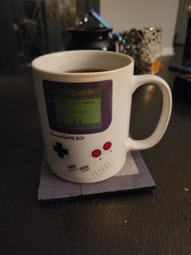 A white mug designed to look like a Game Boy, featuring a small screen and red buttons, sitting on a coaster next to a coffee cup.