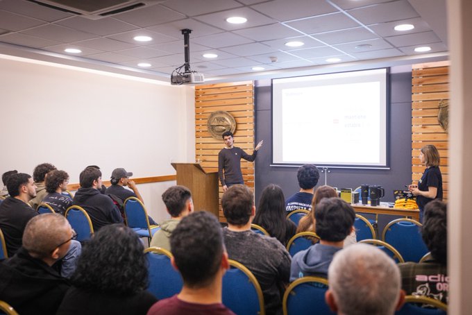 A speaker stands at a wooden podium in a classroom, gesturing toward a projection screen displaying the Binance logo, while addressing an audience of students at Universidad del Aconcagua in Mendoza, Argentina, as part of the Binance Academy University Tour engaging 60 computer engineering students in discussions about blockchain technology.