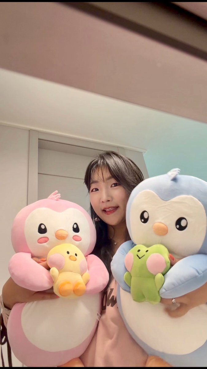 A young woman smiles while holding up two large Rowlet penguin plushies—one pink holding a small yellow duck plush, and one blue holding a green Froakie plush.