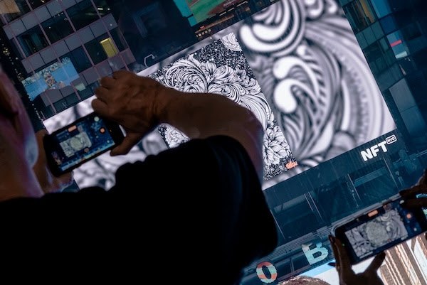 An over-the-shoulder view of a person photographing artwork displayed on a large screen marked "NFT" using a smartphone, with intricate black and white patterns visible on the display.