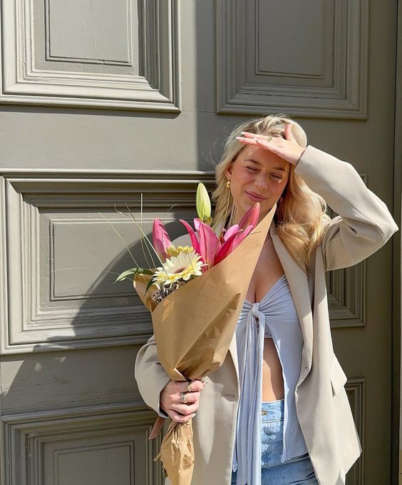 A young blonde woman standing in front of a paneled door, smiling and shielding her eyes from the sun while holding a bouquet of flowers wrapped in brown paper.