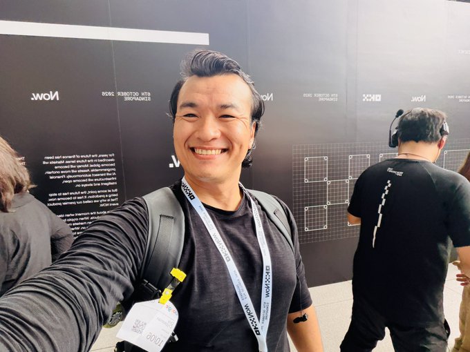 A selfie of a smiling man wearing a black shirt and lanyard in front of a black MoMA wall, with another person in a black shirt standing nearby in what appears to be a design exhibition space.