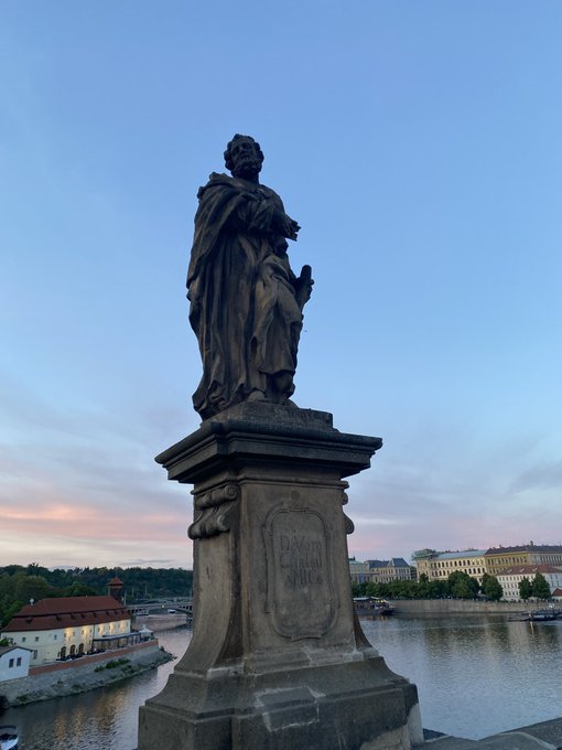A stone statue of a robed figure on a pedestal overlooking a river at dusk, with buildings and a bridge visible in the background, likely the Charles Bridge in Prague.