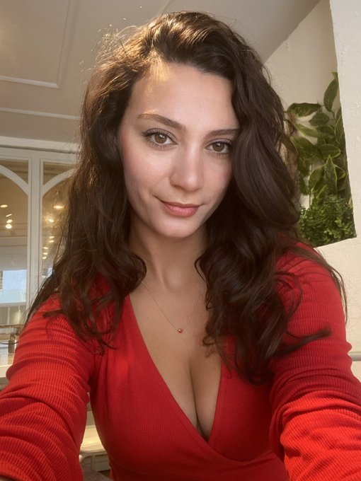 A woman with wavy brown hair smiles at the camera while wearing a red v-neck top and a small heart necklace.