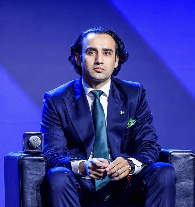 A man with wavy dark hair, wearing a navy blue pinstripe suit with a teal tie and a Pakistani flag pin on his lapel, sits in a dark chair against a blue-lit backdrop. He appears to be at a formal panel or conference, with a microphone visible in the foreground, fitting the context of discussing Pakistan's efficient crypto regulatory framework.