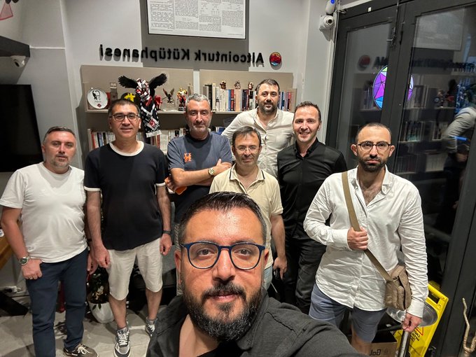 A group selfie of seven men indoors in front of a bookshelf with 'AI Accountancy' signage displayed in reverse, suggesting they are facing a window or mirror.