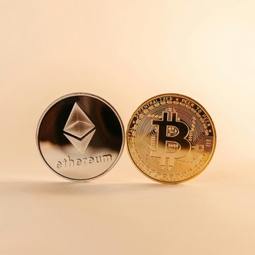 The image shows a close-up view of two coins - one gold coin representing Bitcoin and another silver coin with an "Ethereum" label. They are placed side by side against a light pink background, creating a striking contrast between their metallic colors and the soft gradient hue of the backdrop. The image seems to depict a discussion or analysis about the recent market trends in the crypto space, particularly Bitcoin's and Ethereum's performance.