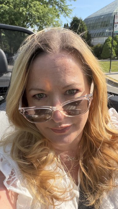The image features a woman wearing sunglasses, with her face turned towards the camera. She has blonde hair that falls over her shoulders, and she is dressed in a white shirt. The background of the photo shows an outdoor setting, possibly a parking lot or street scene. There are also some cars visible in the background.