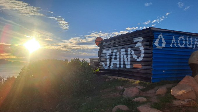 A sunset scene with a sunburst on the left and a blue wooden container on the right featuring graffiti that reads "JAWS" and "AQUA" on a rocky landscape.