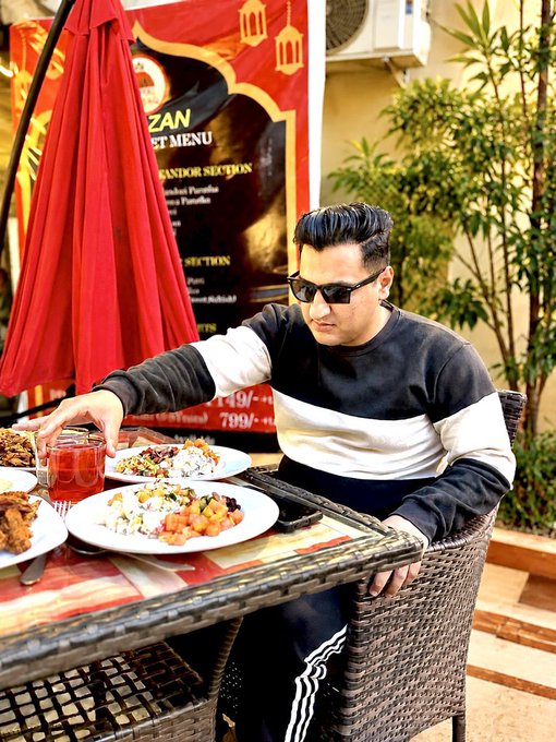 A man wearing sunglasses and a black-and-white striped sweater sits at an outdoor restaurant table with plates of food and a drink in front of him, with a menu displaying prices like "149/-" and "799/-" visible behind him. The post references preparing for a day of trading, and the image metaphorically represents "fueling up" with a hearty meal before tackling the markets—serving as a power-up moment before opening multiple trades.