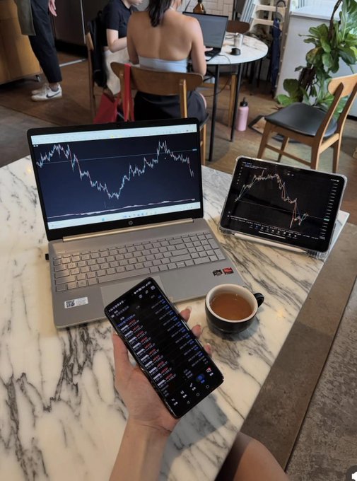 A first-person view of someone holding a smartphone displaying stock market data, with a laptop and tablet also showing trading charts on a marble cafe table next to a cup of coffee.
