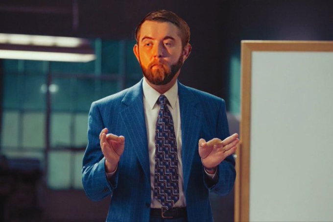 A man with a beard wearing a blue suit and patterned tie stands in an office setting with his hands raised in a gesturing motion.