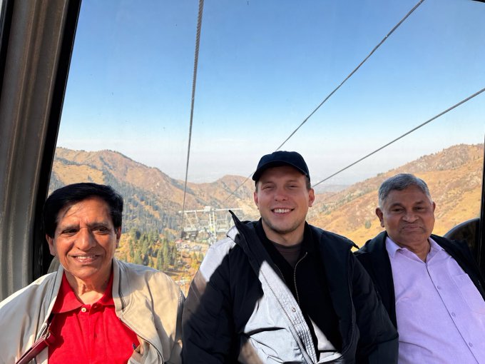 Three smiling men posing for a photo inside a cable car with mountain views and a valley town visible in the background.
