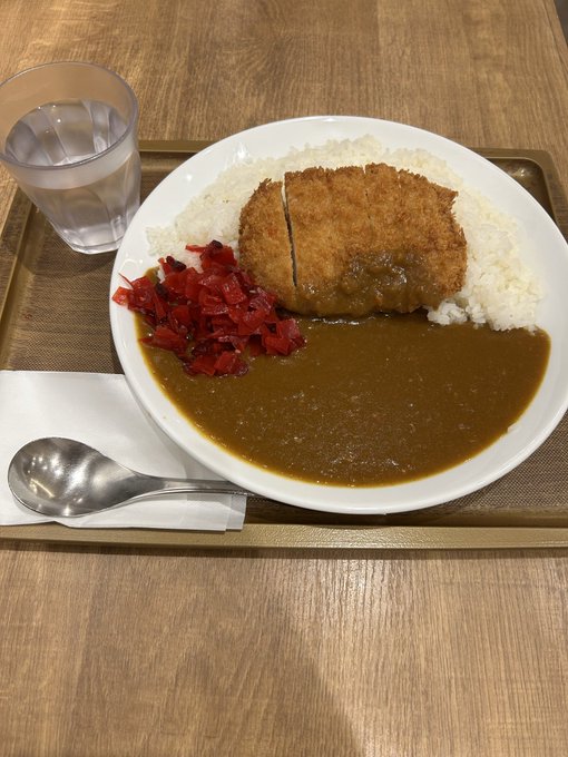 A Japanese curry rice plate served on a tray with a breaded cutlet, fukujinzuke pickles, and a glass of water on a wooden table.