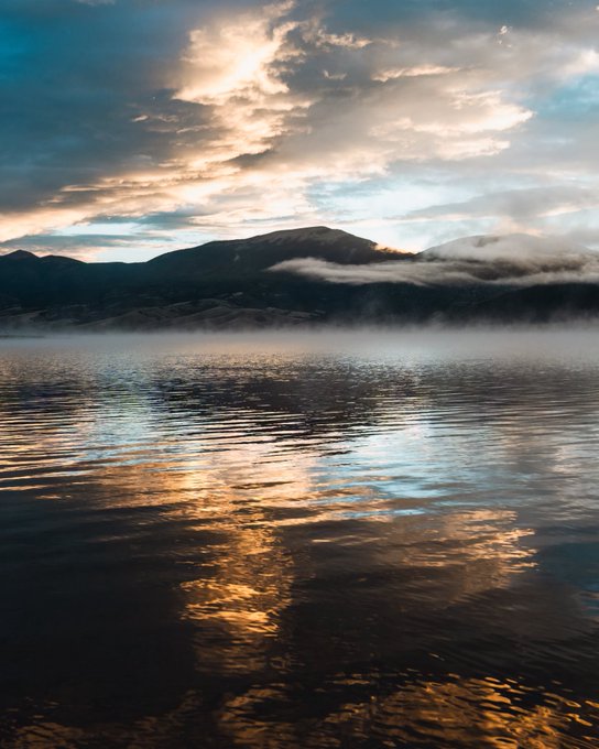 A scenic landscape photograph of a mountain range with clouds and mist reflected on a calm body of water at sunset or sunrise.
