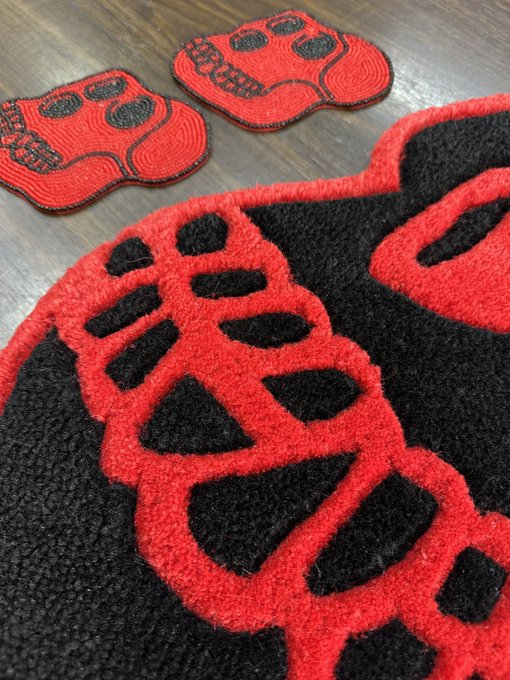 Close-up photograph of red and black skull-themed textile items, including a doormat and smaller patches, resting on a wooden floor.