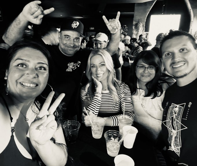 A black and white selfie of five smiling friends posing at a bar with drinks on the table, throwing up rock-and-roll hand gestures to celebrate the "Mutant Apes" 5-year anniversary as the group enjoys a night out together.