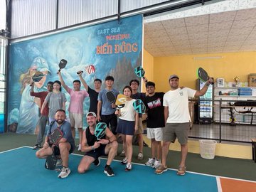 The image shows a group of people posing for a photo with their paddle sports equipment, including tennis rackets and paddleboards. The group consists of at least 12 individuals who are standing together against a backdrop featuring a mural or painting. The scene appears to be set in an indoor space, possibly a gymnasium or a similar venue where various activities take place.