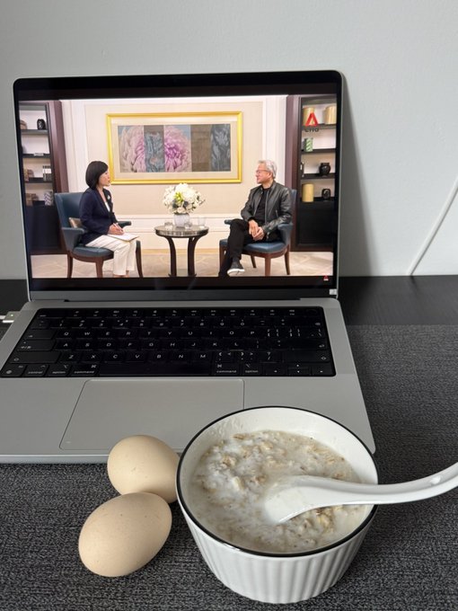 A breakfast scene featuring a bowl of oatmeal with a spoon and two brown eggs placed in front of an open laptop playing a video of two people being interviewed.