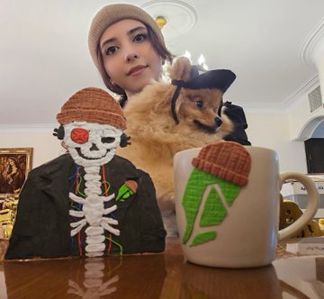 A woman poses behind a cardboard art piece of a skeleton character wearing a beanie and a furry cap, alongside a matching decorated mug featuring the same character, with a small dog perched between them. The artwork is a 3D-textured creation of the "@crypto_czar_eth" profile, made with air-dried clay on cardboard as mentioned in the post.
