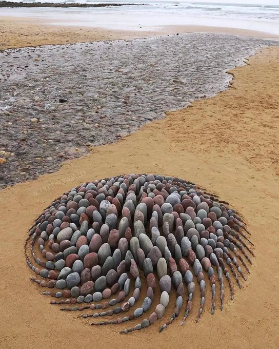 A circular land art arrangement of smooth, multicolored pebbles placed on a sandy beach near the shoreline, attributed to the artist Soul in World.