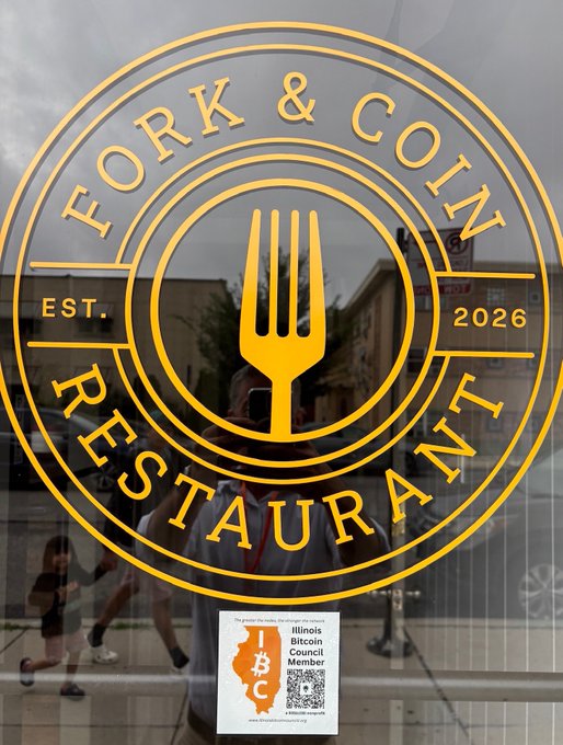 The window of a restaurant displaying a gold circular logo with a fork that reads 'FORK & COIN RESTAURANT EST. 2026'. An Illinois Bitcoin Council member sticker is visible on the window, along with reflections of the photographer and street.