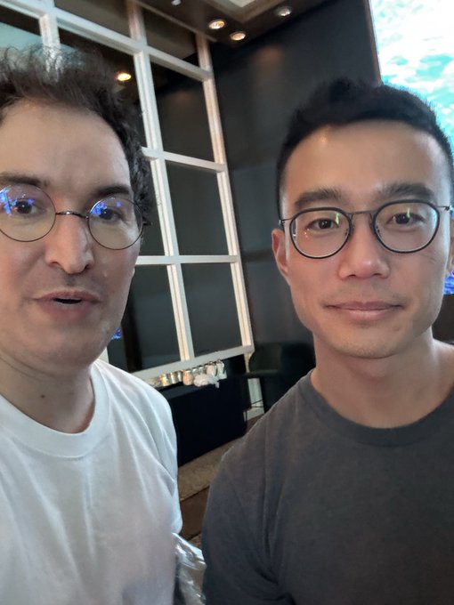 A selfie of two young men wearing glasses indoors, one in a white t-shirt and the other in a gray t-shirt, with a window and dark paneled wall behind them.