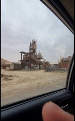 A view through a car window of a construction or industrial site with cranes, scaffolding, and dirt mounds under an overcast sky.