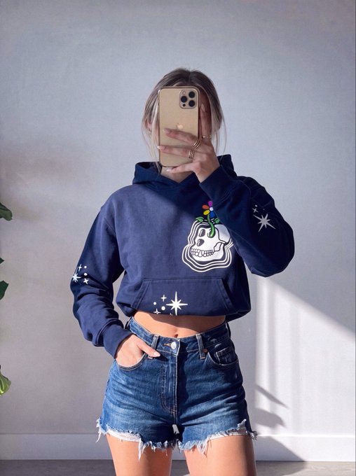 A person takes a mirror selfie wearing a navy blue hoodie featuring a skull graphic with a flower, paired with distressed denim shorts.