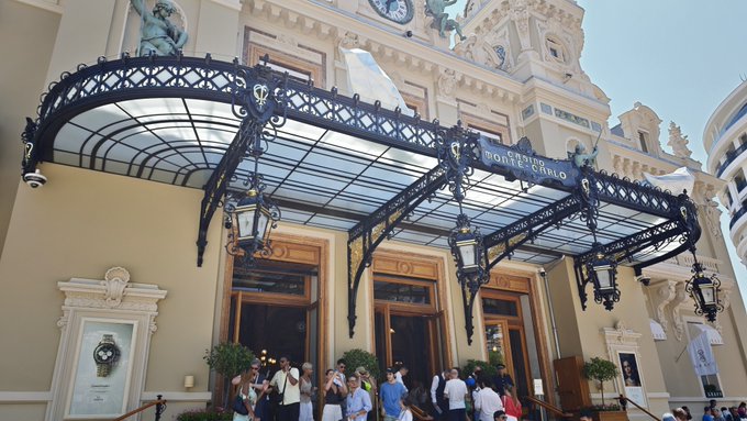 Photo of the ornate entrance of the Casino de Monte-Carlo in Monaco, featuring an elaborate wrought-iron and glass canopy with statues, and crowds of tourists gathered below.