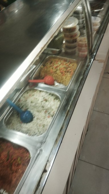 A blurry photo of a food service counter showing metal trays containing different dishes, including rice and noodles, with serving utensils.