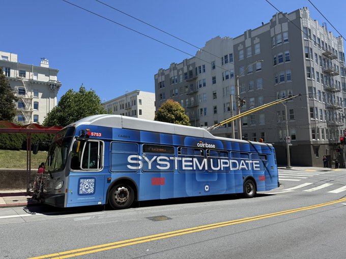 The image features a blue bus parked at a bus stop, with a person standing nearby. A sign on the side of the bus reads "System Update". The background shows buildings, indicating that this scene takes place in an urban setting.