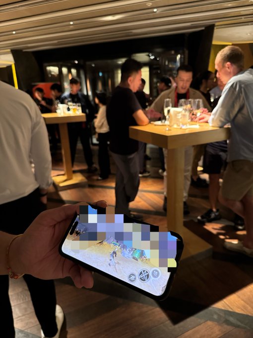 A hand holds a smartphone displaying a pixelated game interface in the foreground, while a social gathering of people stands around high-top tables in a dimly lit venue, with wine glasses visible on the tables, illustrating a post-event networking scene at a crypto conference discussing market fallout.