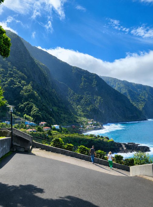 A breathtaking morning greeting from a scenic coastal overlook, where two people pause to admire lush green cliffs plunging into a turquoise sea under a bright blue sky — perfectly matching the cheerful "gm to absolutely everyone" vibe with its sunny, uplifting start to the day.