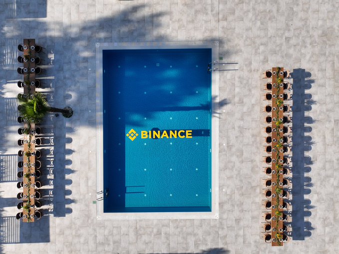 The Binance logo is displayed at the bottom of a swimming pool, captured from an aerial view, with rows of lounge chairs lined up on both sides, fitting the post's "GM" (Good Morning) greeting.