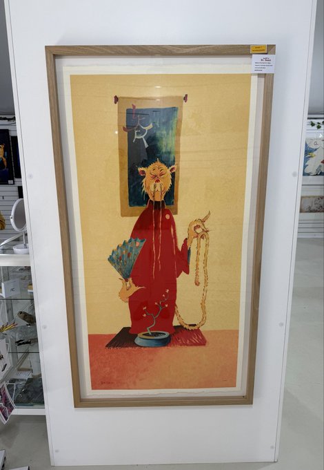 A framed traditional East Asian-style painting depicting an anthropomorphic cat figure in a red robe holding prayer beads and a peacock feather fan, hanging in what appears to be a shop or gallery.