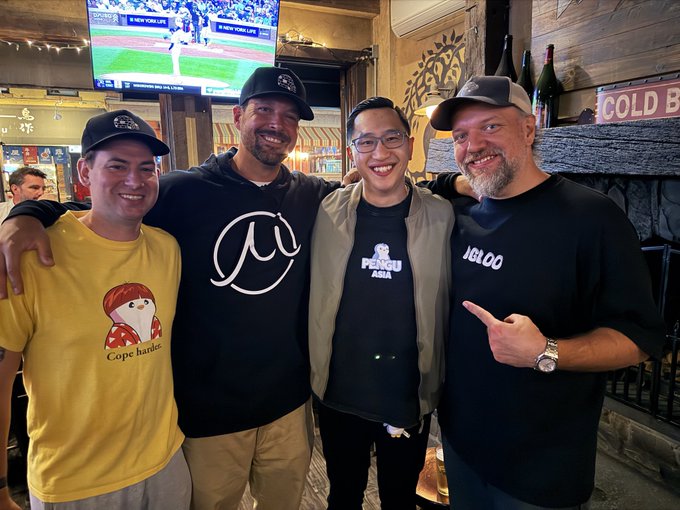 Four smiling men pose together for a photo in what appears to be a bar or restaurant with TVs and a "COLD B" sign visible in the background; one wears a yellow "Cope harder" shirt, another a black hoodie with a white "@" logo, another a "PENGU ASIA" shirt, and the last a black "IGLOO" t-shirt.