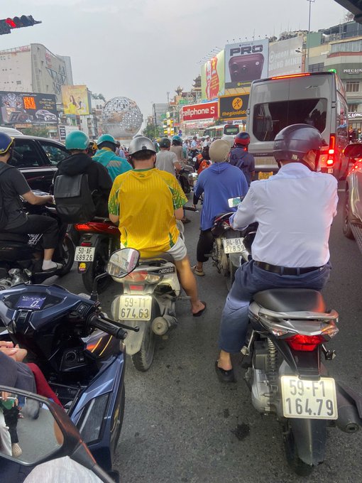 A bustling traffic scene filled with motorcyclists and scooter riders, many wearing helmets and face masks, stopped at a light in a busy urban area with billboards visible in the background.