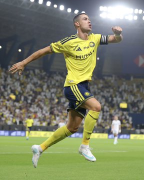 A soccer player in a yellow Fenerbahçe kit celebrating on the field with arms outstretched during a match in front of a stadium crowd.