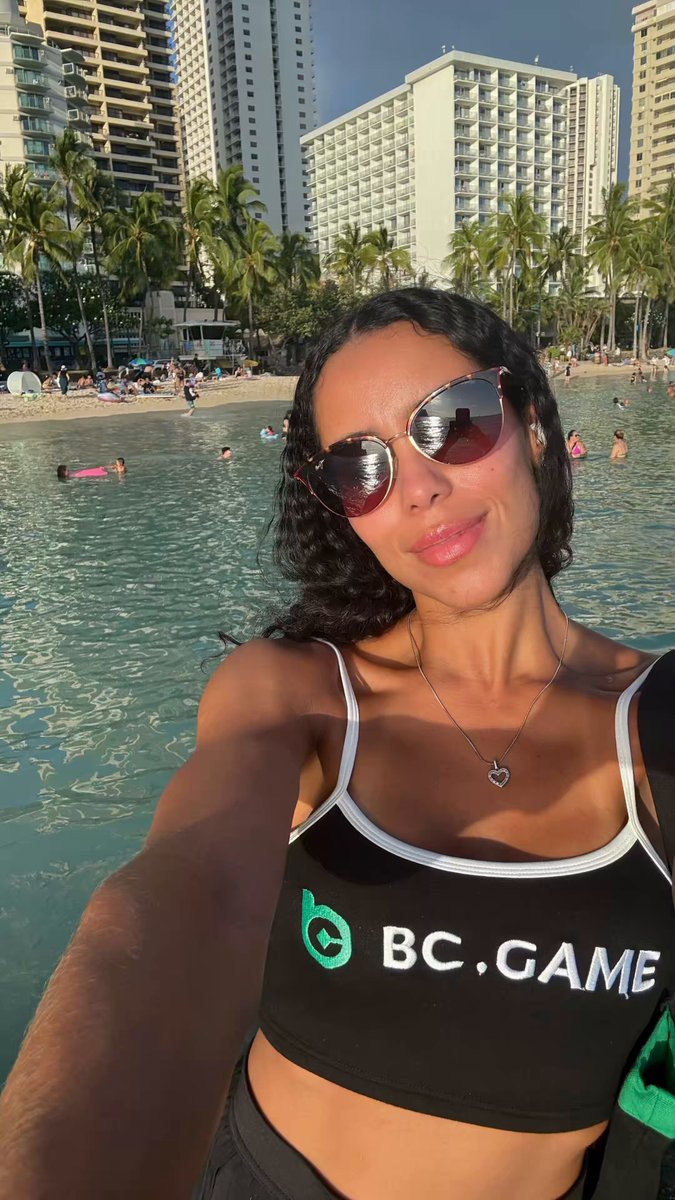 The image features a woman wearing a black tank top, sunglasses, and a necklace. She is standing by the water at the beach, taking a selfie using her phone's camera. The photo captures the beautiful sunset over the ocean, creating a serene atmosphere for the woman as she poses with her phone in hand.