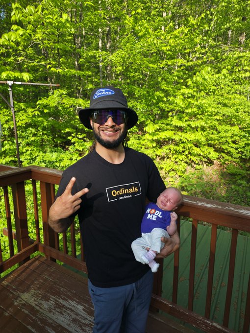 The image features a man holding his baby, both smiling for the camera while standing on a wooden deck or porch. The man is wearing sunglasses and a hat, giving him a cool and relaxed appearance. In the background, there are trees that add to the outdoor setting of the scene. The caption "GM Reppin BTC and Ordinals strong. Have a wonderful day y'all!" suggests that this image might be related to a post on social media or a personal message from someone who wants to share their experience with others.