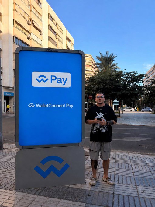 Man in a skull t-shirt standing next to a large WalletConnect Pay advertising display on a city street.